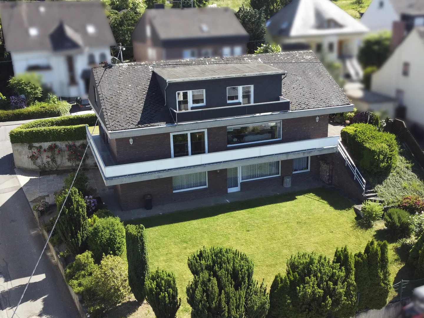 Aerial view of a two-story brick house with a large green lawn, balconies, and surrounding hedges in a suburban area.