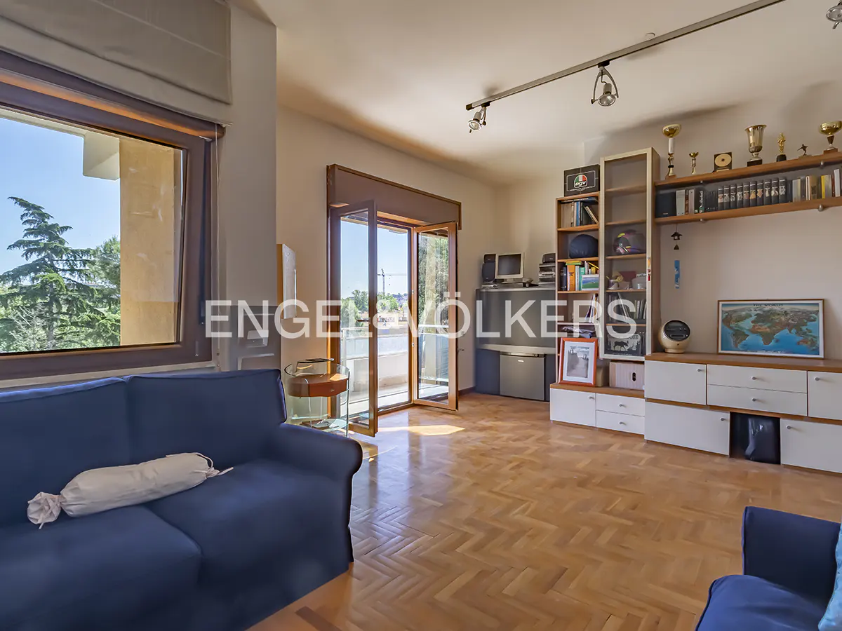 A bright living room with a blue sofa, wooden floors, and a balcony with an open door. A bookshelf and a TV stand are against the wall.