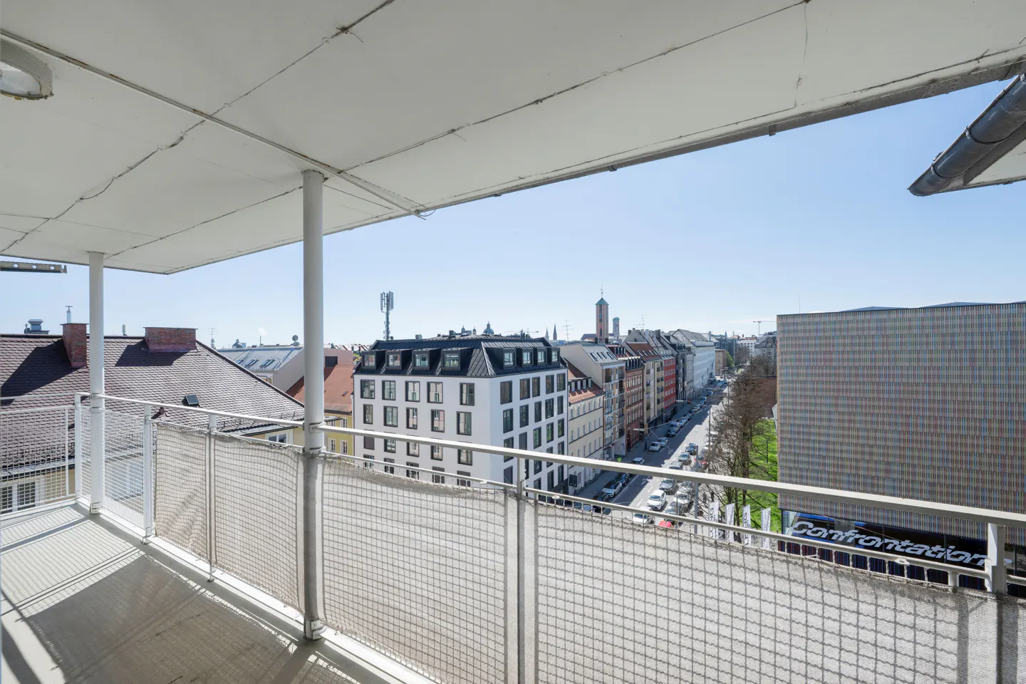 View from a balcony with white metal railings overlooking a city street with buildings and cars on a sunny day.