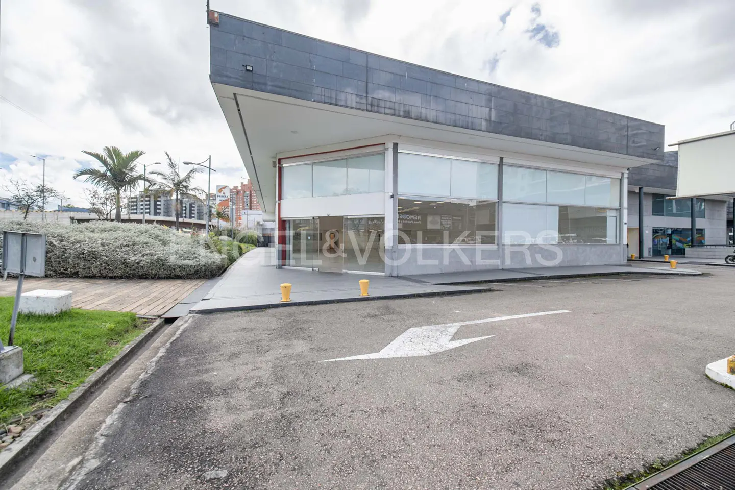 Engel & Volkers office building with large windows and a gray facade, set against a cloudy sky. A white arrow is painted on the asphalt.