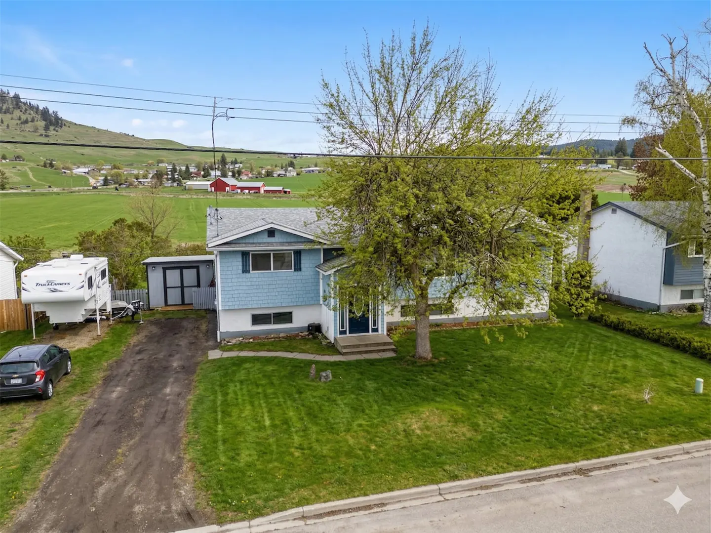 Two-story blue house with a green lawn and a large tree in the front yard. A car and camper are parked in the driveway. Rolling green hills in the background.