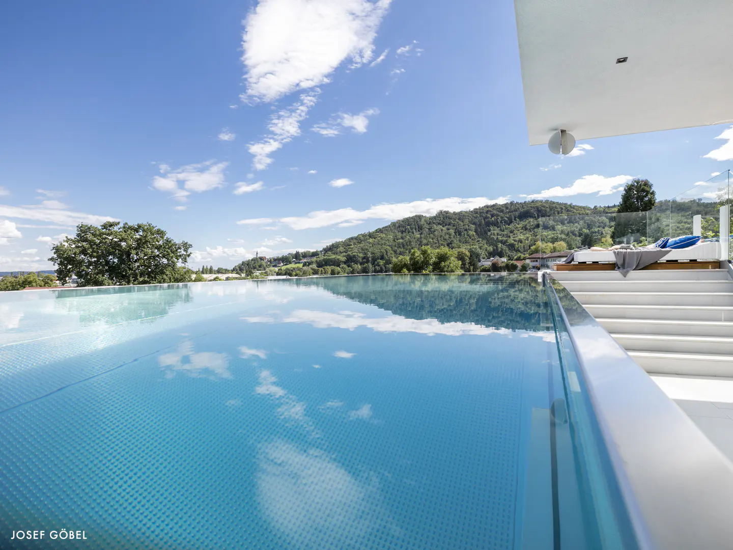 Infinity pool with clear blue water reflecting the sky, overlooking a lush green mountain landscape on a sunny day.