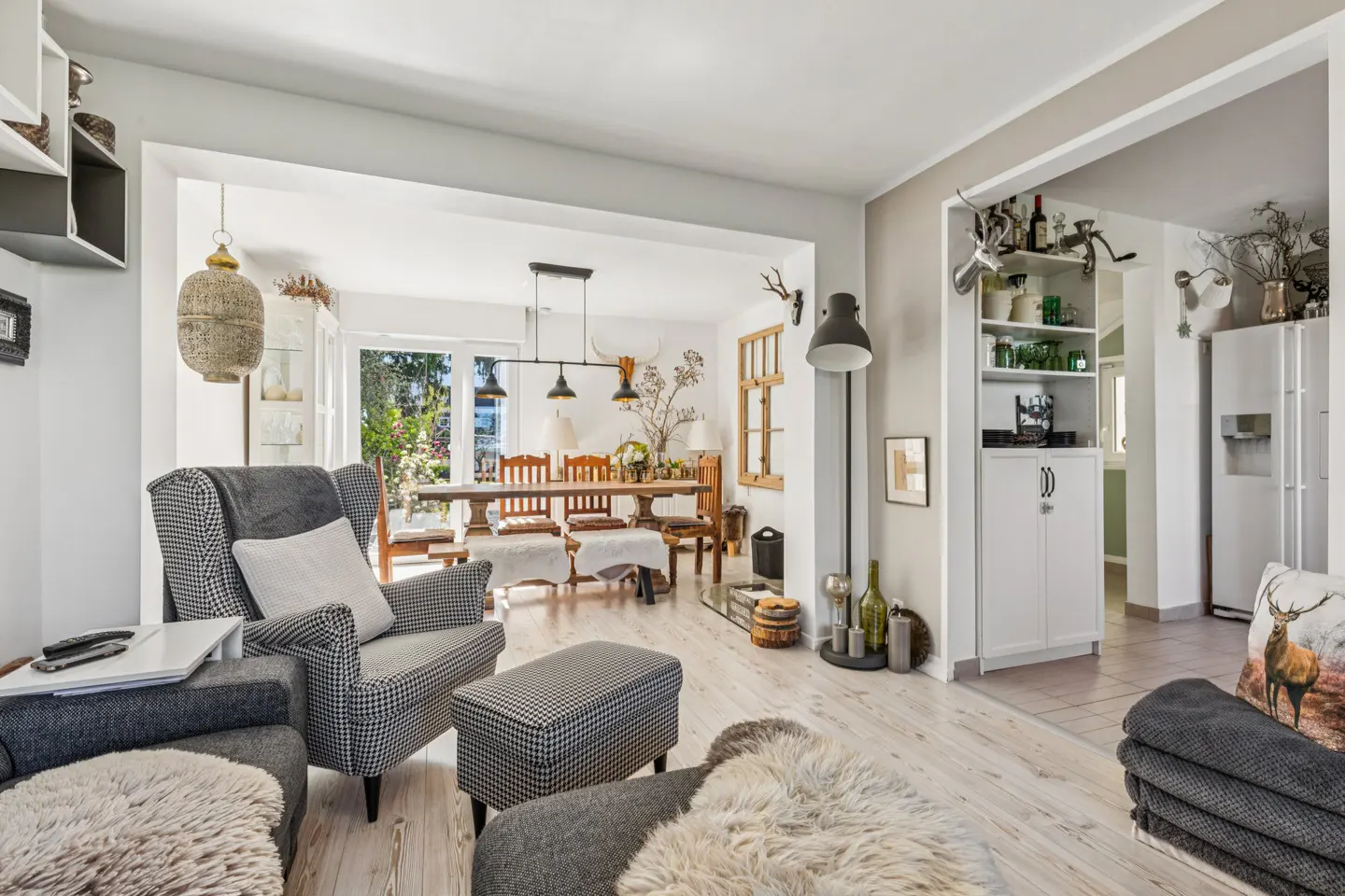 A bright, open-concept living and dining area with light wood floors. A patterned armchair and ottoman are in the foreground. A dining table is visible through an archway.