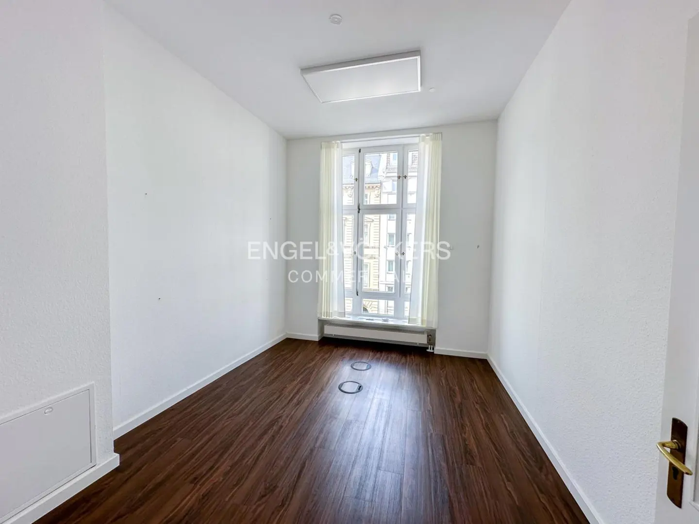 Bright, empty room with dark wood floors, white walls, and a large window with sheer curtains. A rectangular light fixture is on the ceiling.