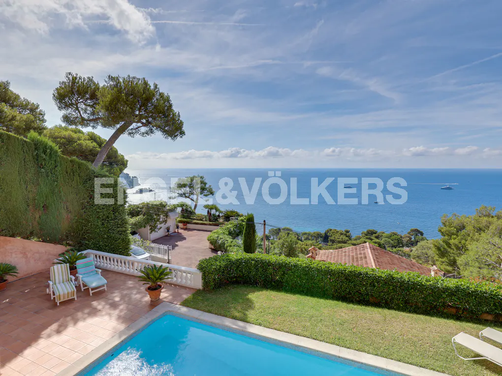 A sunny terrace with a swimming pool, lounge chairs, and lush greenery overlooking a vast blue sea under a clear sky.