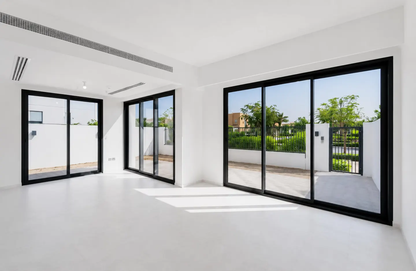 Bright, empty room with white walls, polished floors, and large black-framed sliding doors opening to a garden patio.