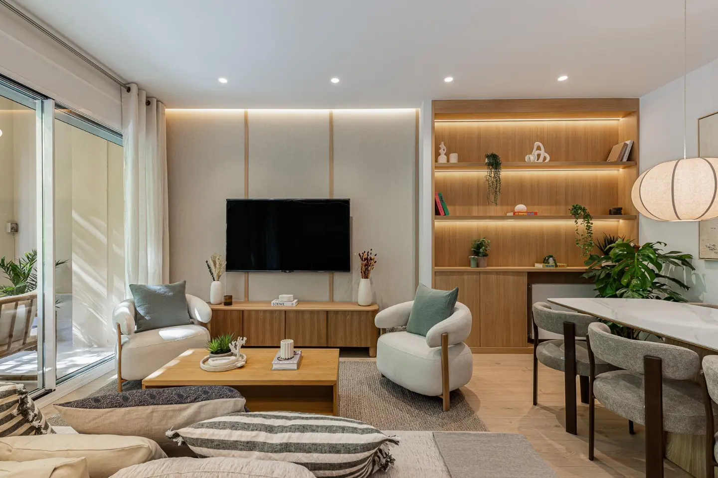 Modern living room with a wall-mounted TV, wooden media console, two cream armchairs, and a dining area with wood shelving.