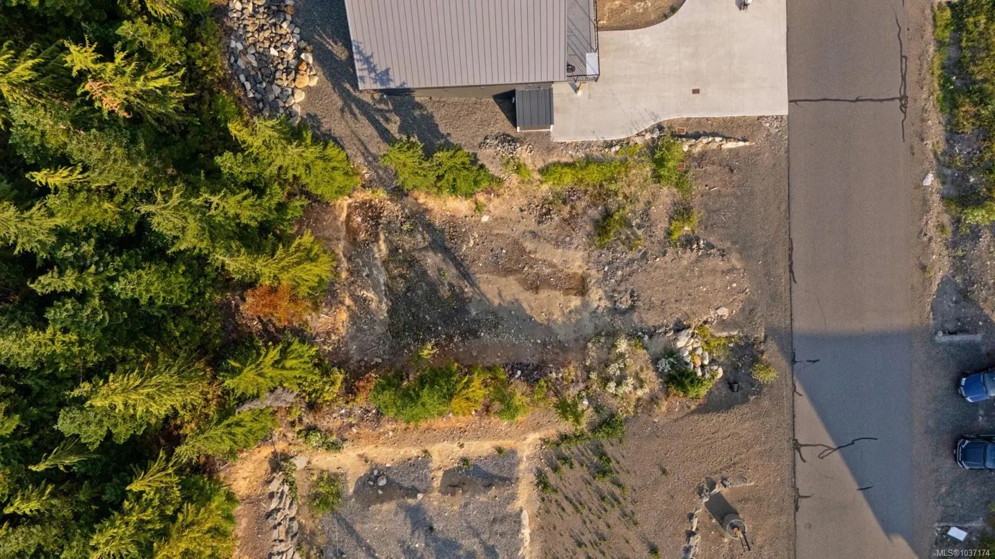 Aerial view of a vacant lot next to a house and a road, with trees on one side. The lot is mostly dirt and gravel, with some small plants growing.