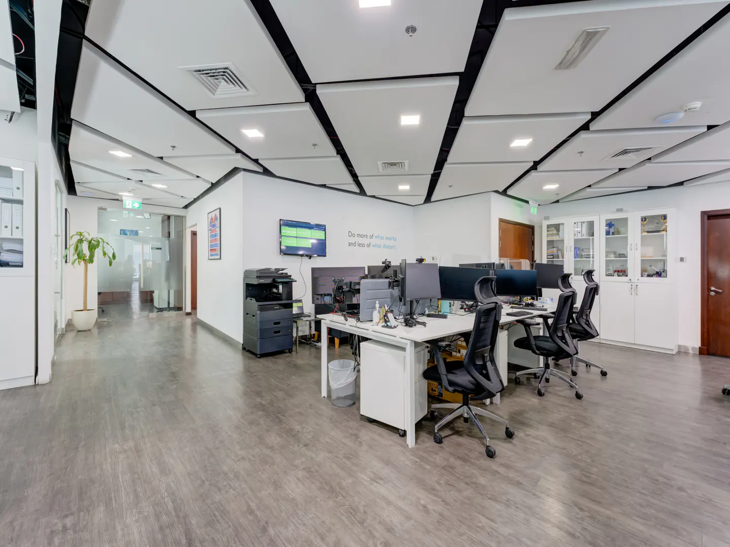 Bright office space with white walls, gray wood-look floors, and modern ceiling panels. Desks with computers and black chairs are arranged in rows.