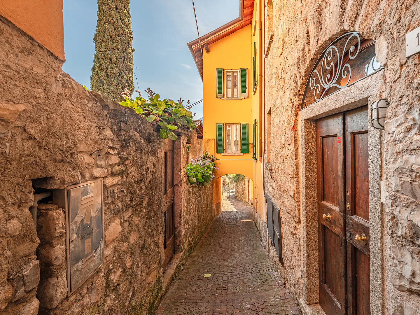 A narrow cobblestone street between stone buildings, one yellow with green shutters, leads to an archway.