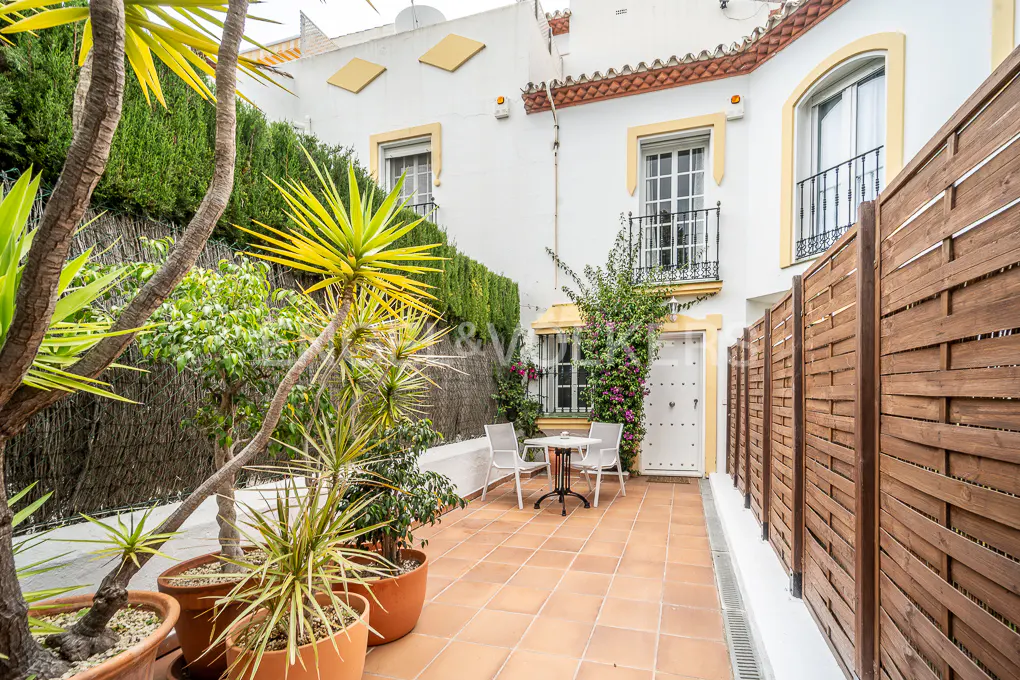 A courtyard with terracotta tiles, a table and chairs, and a white building with yellow trim. Potted plants and a wooden fence add privacy.