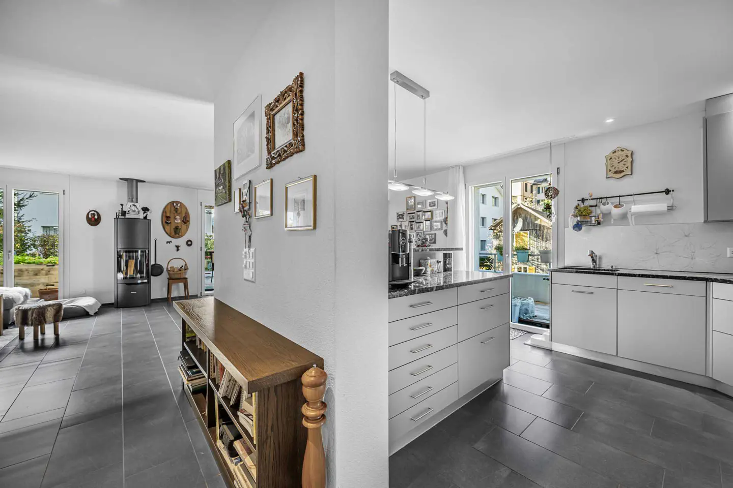 A modern home interior with a living room, hallway, and kitchen. The living room has a wood-burning stove and a window to the outside. The kitchen is white with a marble countertop.