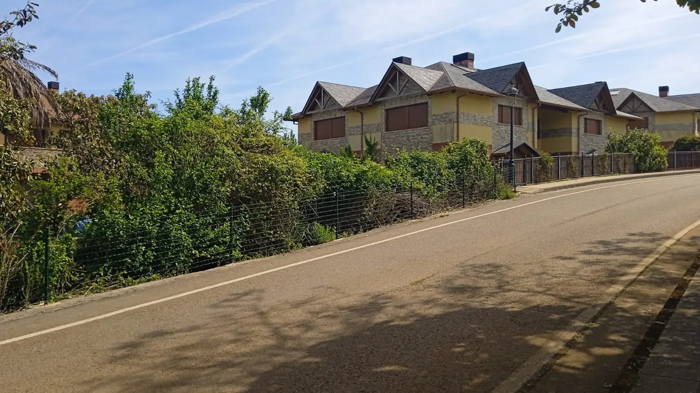 Street view of yellow and stone townhouses with gray roofs behind a green hedge and black fence.