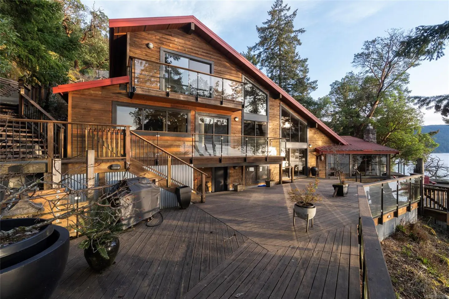 Exterior view of a wood-sided house with a red roof and a large wooden deck overlooking the water.