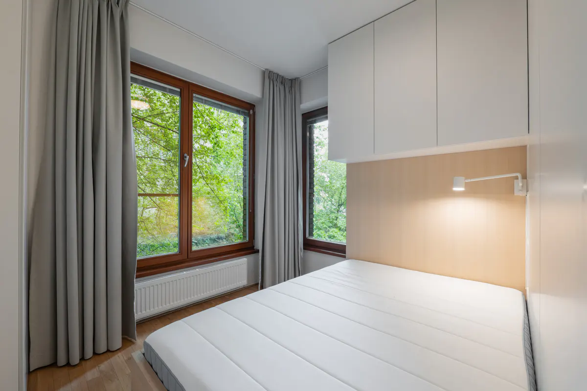 Bedroom with a white mattress bed, wood floors, and two windows with brown frames. Gray curtains hang on either side of the windows, with green trees visible outside.