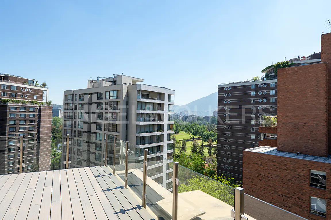 View from a rooftop deck with glass railings, overlooking city buildings and a green golf course on a sunny day.