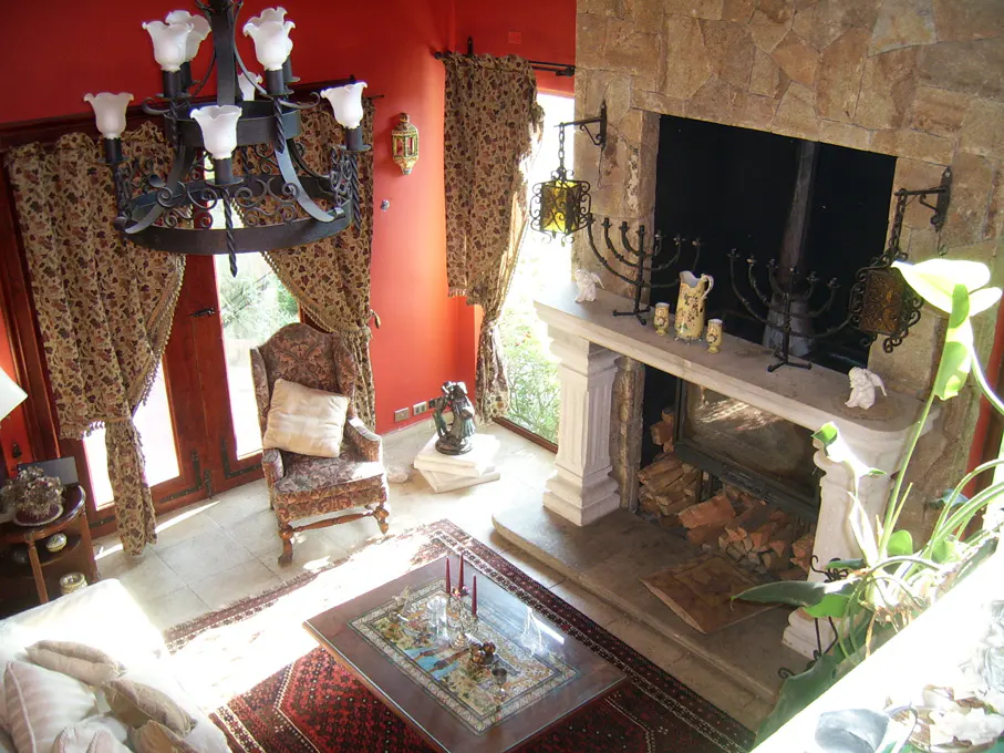 Living room with red walls, stone fireplace, and patterned curtains. A chandelier hangs above a glass-topped coffee table on a red rug.