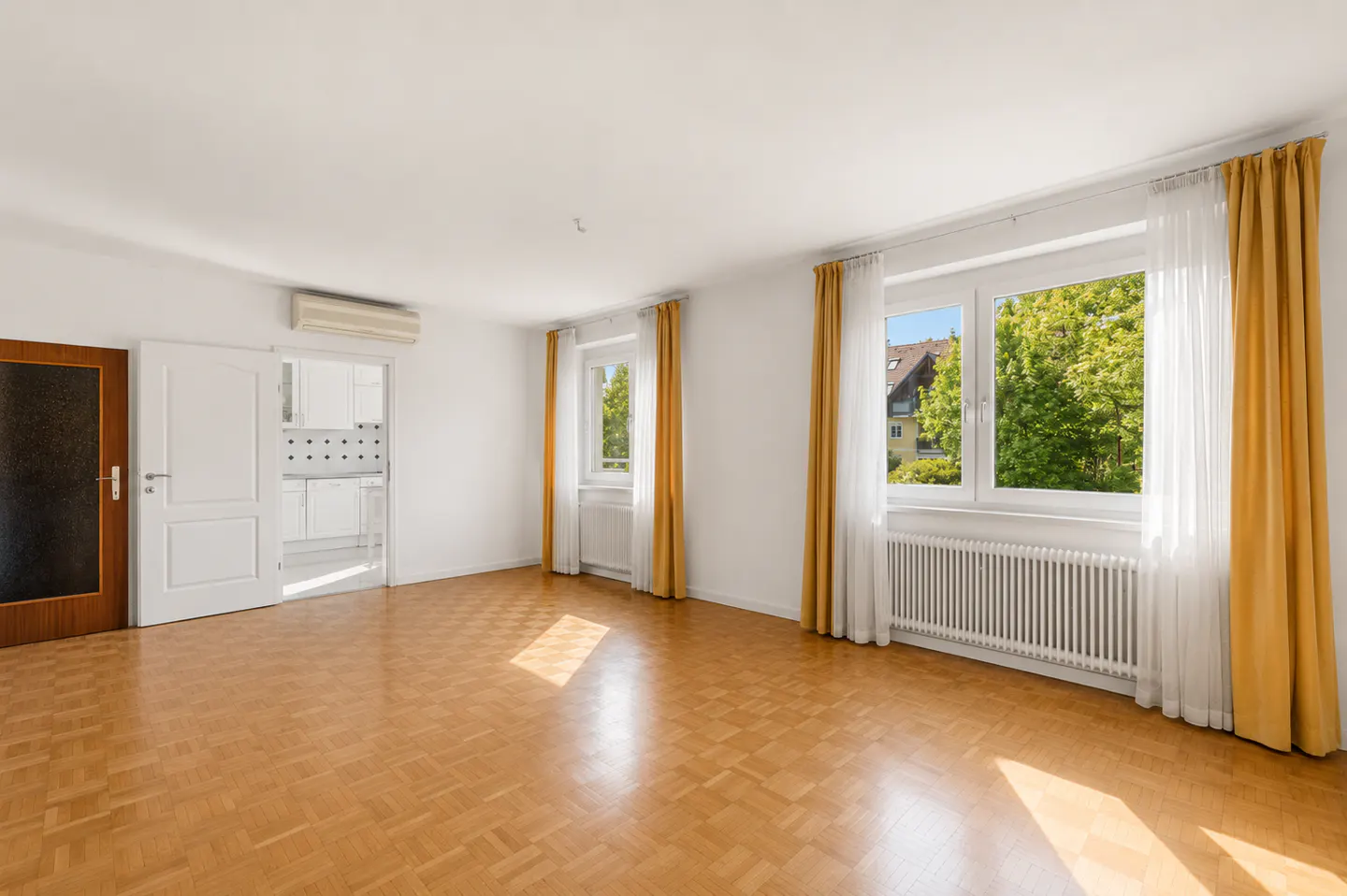 Bright, empty room with parquet floors, white walls, and two windows with yellow curtains. A door leads to a white kitchen.