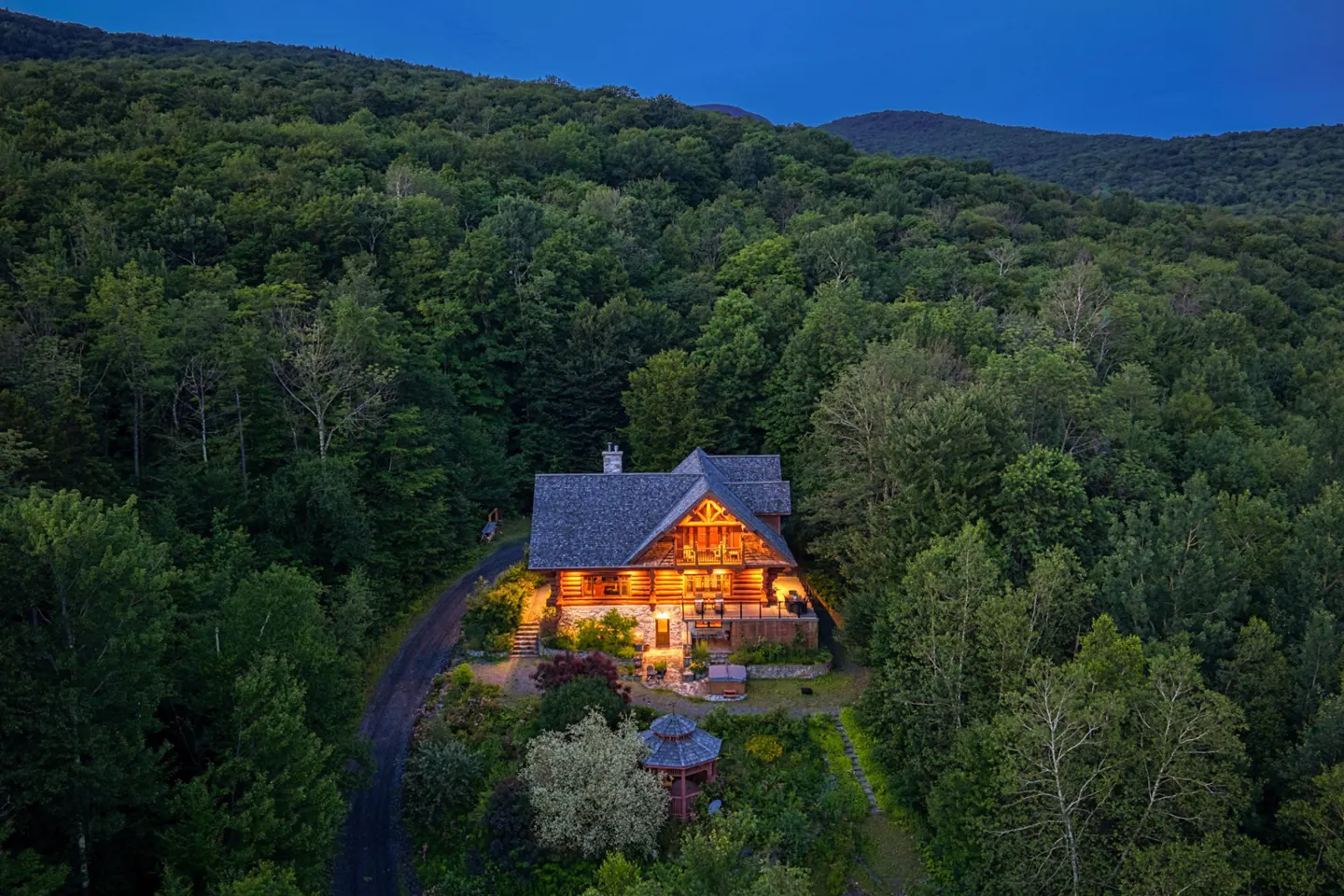 A log cabin glows warmly amidst a dense green forest under a twilight sky.