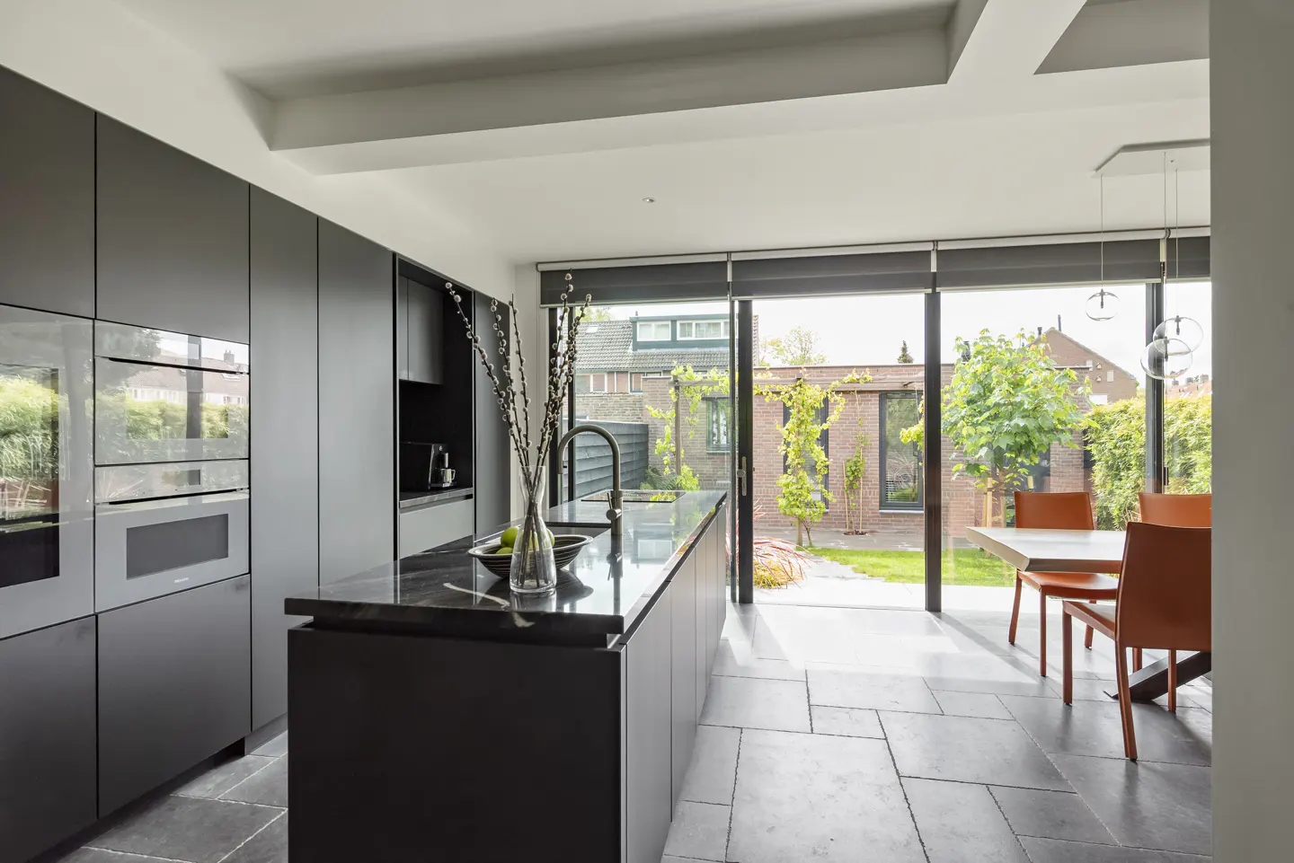 A modern kitchen with dark cabinets, a black countertop island, and a view of a green backyard through sliding glass doors.