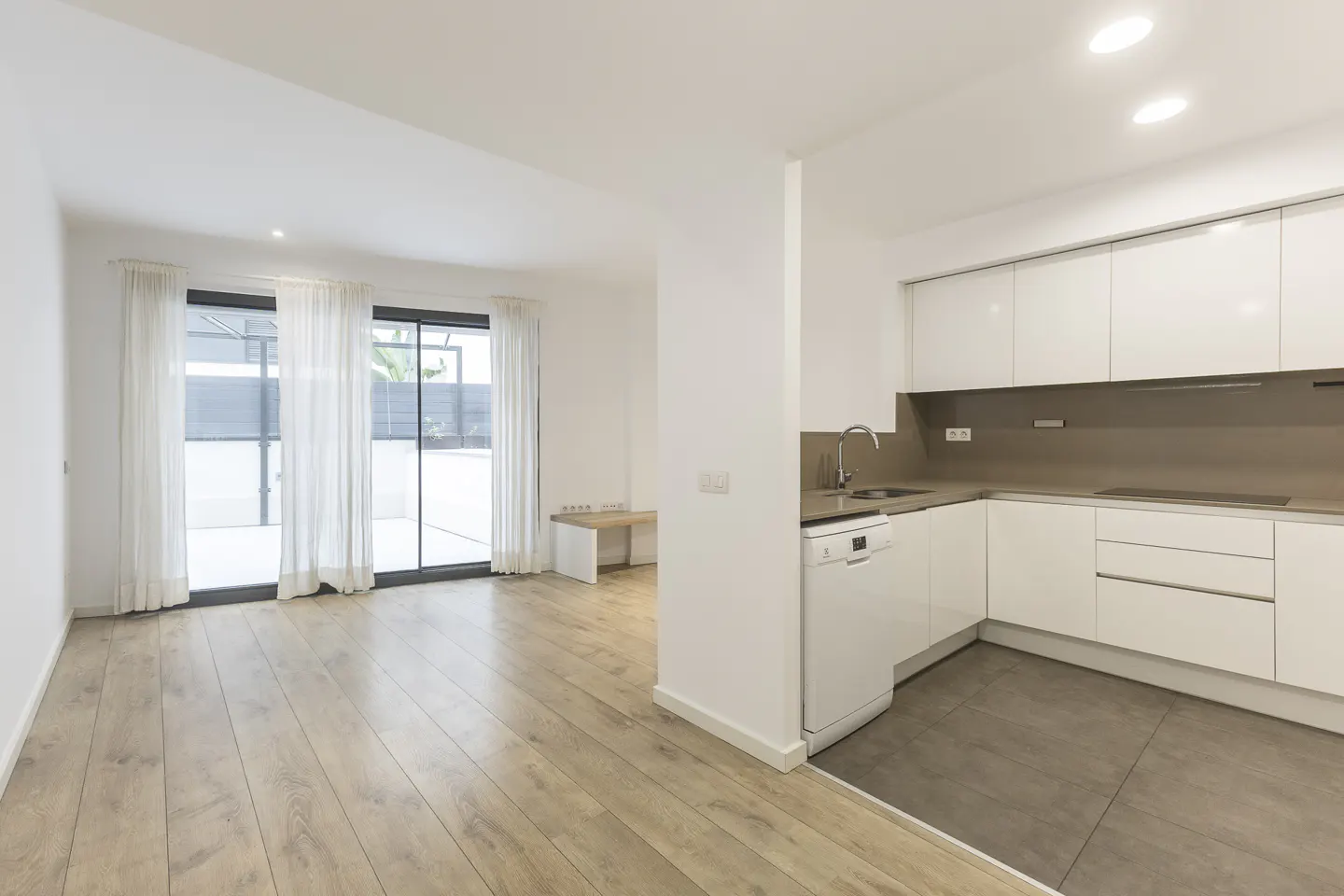 Bright, modern open-plan living space with wood floors, white walls, and a kitchen with white cabinets. Sliding glass doors lead to an outdoor area.