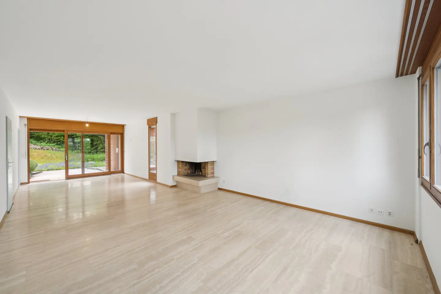 Bright, empty living room with marble floors, white walls, and a brick fireplace. Sliding glass doors lead to a green yard.