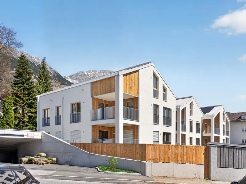 Modern white apartments with wood accents and balconies, set against a backdrop of mountains and a clear blue sky.