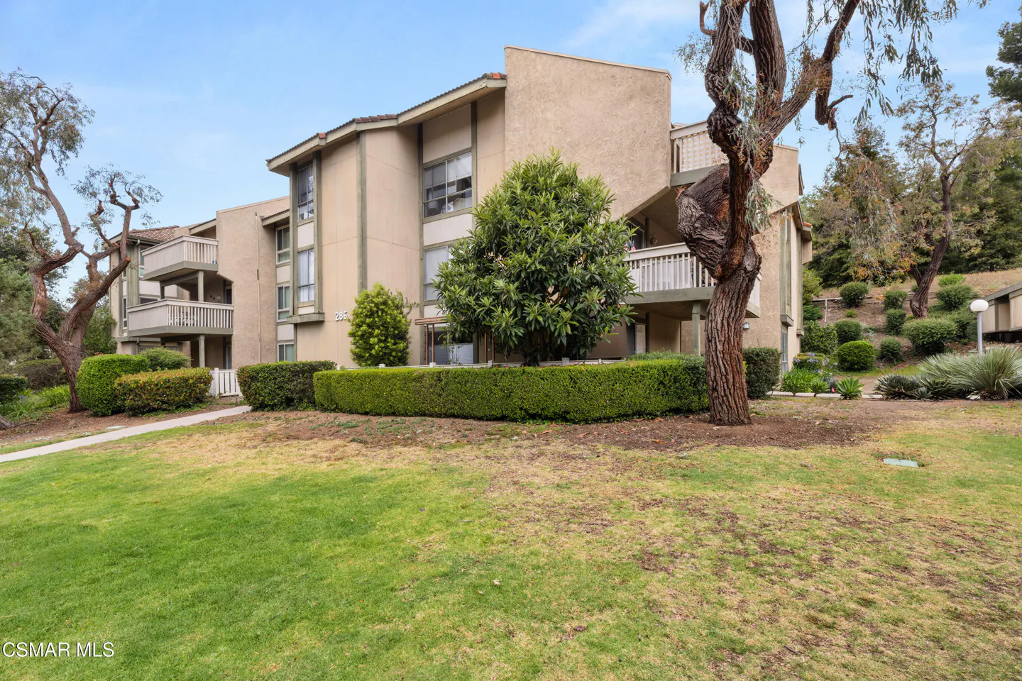 A tan apartment building with white balconies and green trim, surrounded by green grass and trees.
