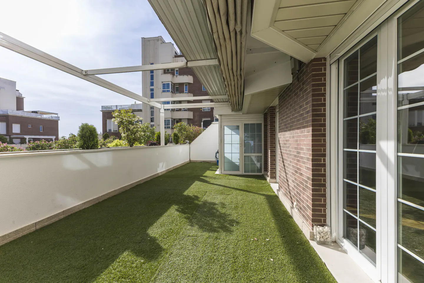 A balcony with artificial grass, a white railing, and a brick wall. White framed glass doors lead inside. A retractable awning provides shade.