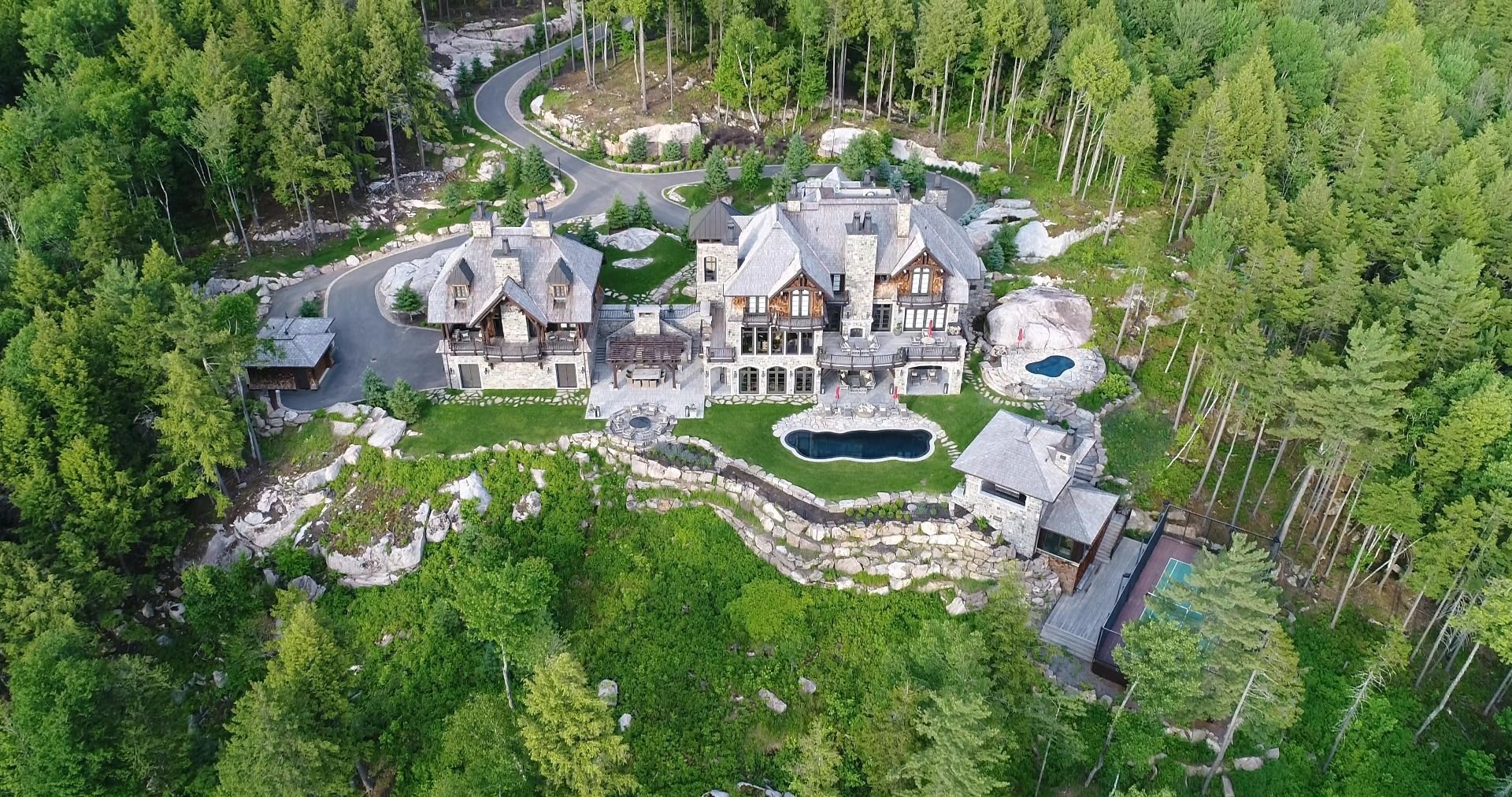 Aerial view of a stone mansion nestled in a lush forest, featuring a swimming pool, tennis court, and winding driveway.