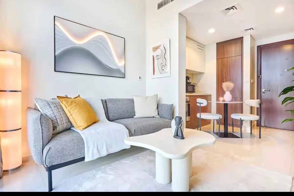 A modern living room with a gray sofa, white rug, and unique coffee table. Artwork adorns the walls, and a small dining area is visible in the background.