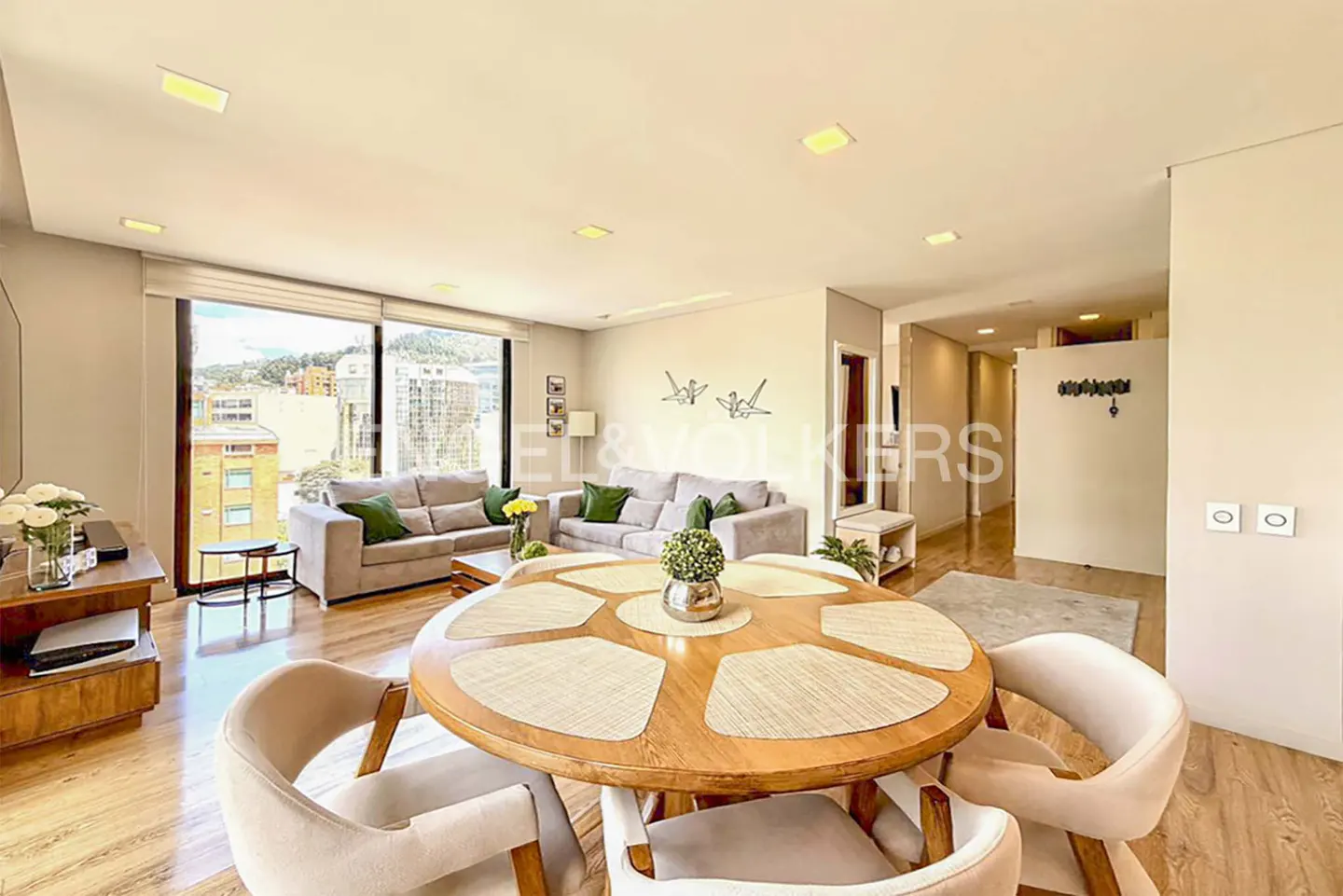 Bright living room with a round wooden table, beige chairs, sofas, and a large window with a city view.