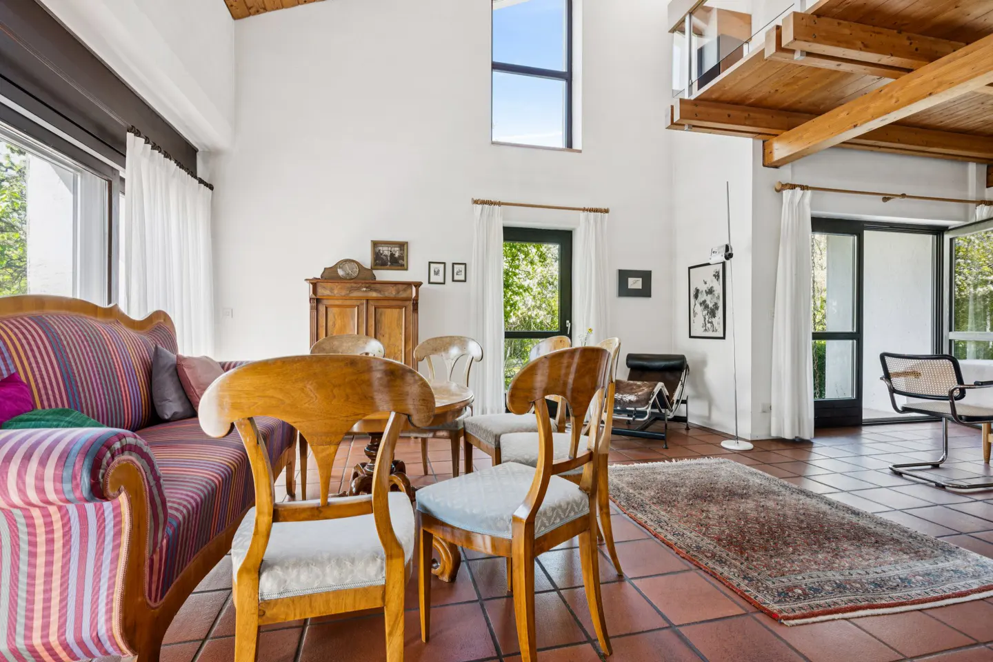 Bright living room with high ceilings, terracotta tile floor, and wooden beams. A striped sofa and chairs surround a rug. Doors lead to the outside.