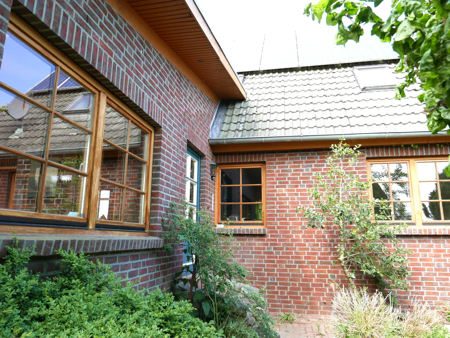Exterior view of a red brick house with wood-framed windows and a gray tiled roof. Green bushes are in the foreground.