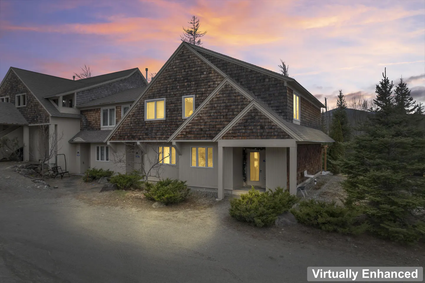 A two-story townhouse with brown shingle siding and a beige stucco base at dusk. Windows glow with warm light. Evergreen trees surround the property.