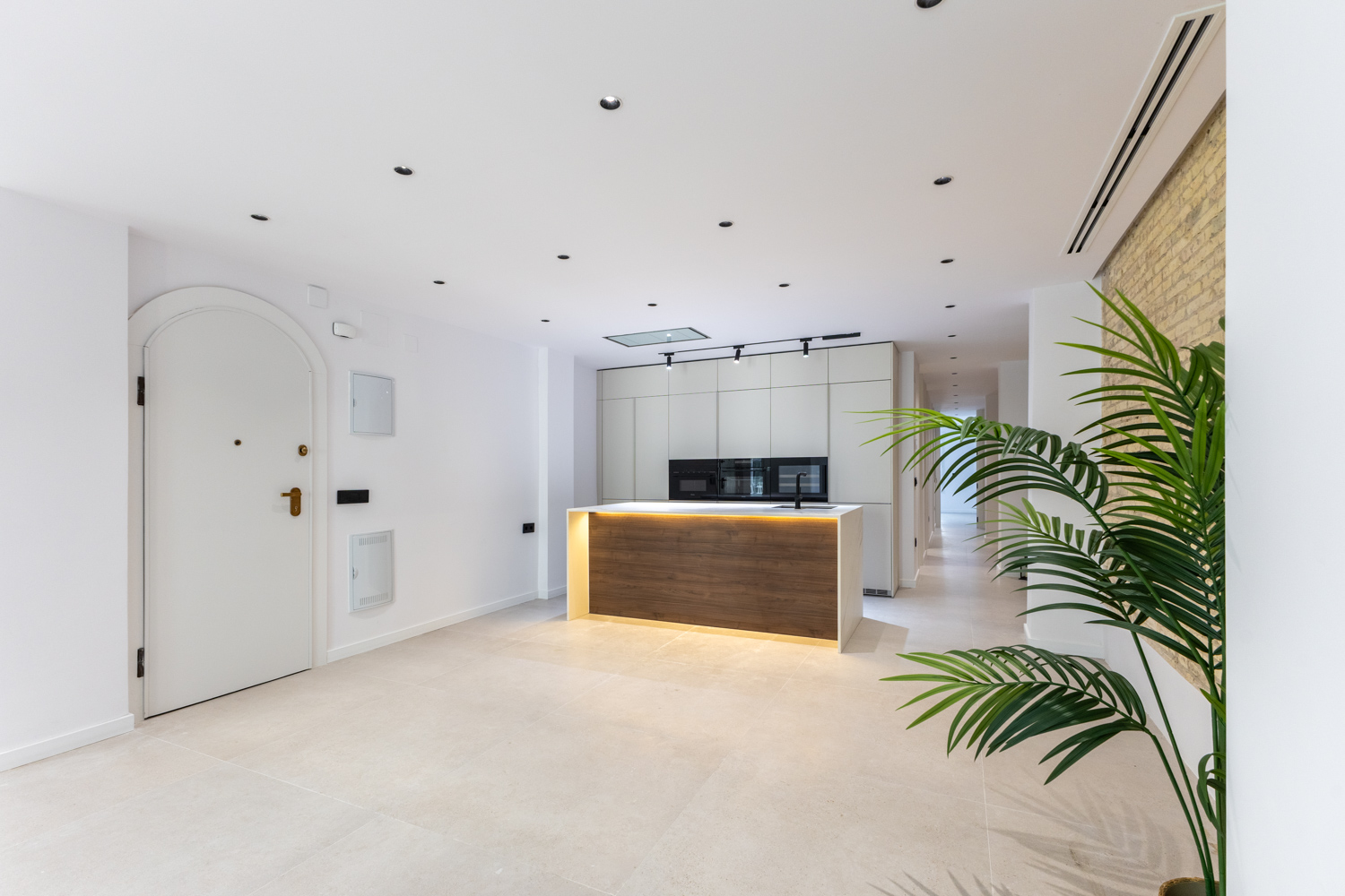 Modern kitchen with a wood-paneled island, white cabinets, arched door, and a large potted plant against a brick wall. [AI-generated text]