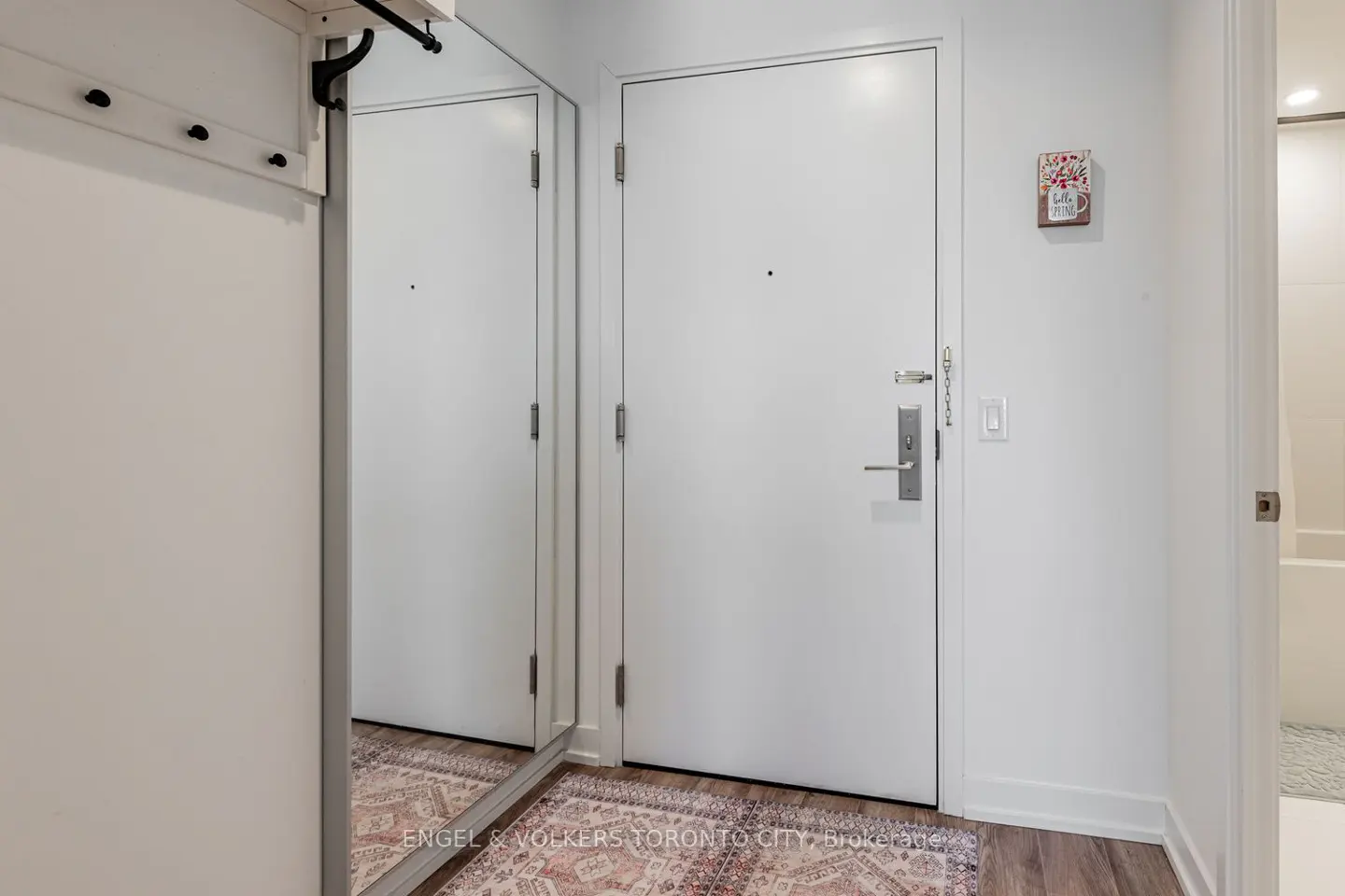 White entryway with a mirror, a coat rack, a white door with a peephole, and a patterned rug on a wood floor.
