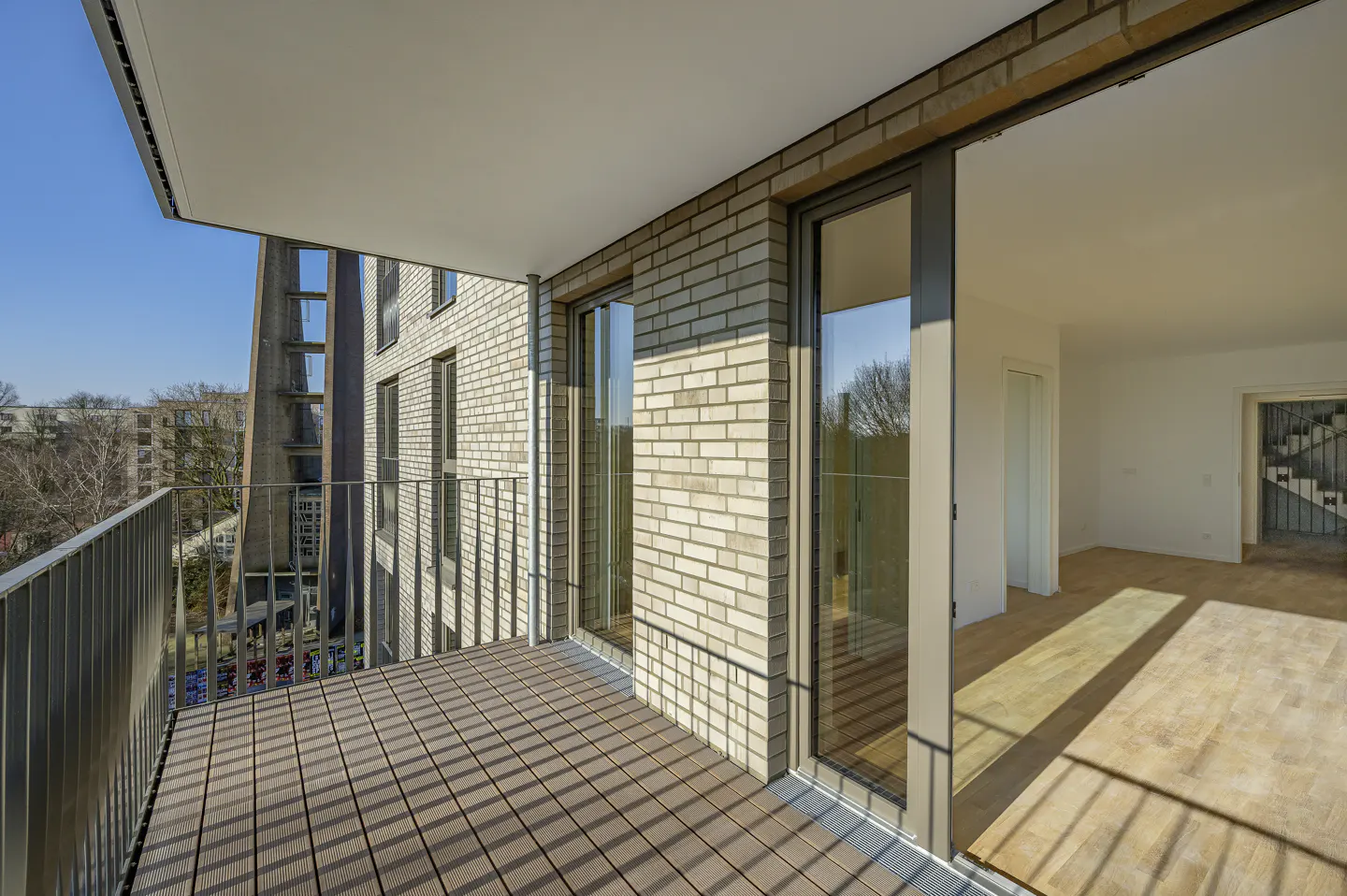 Modern balcony with wood-look decking and metal railing, overlooking a brick building and trees under a clear blue sky.