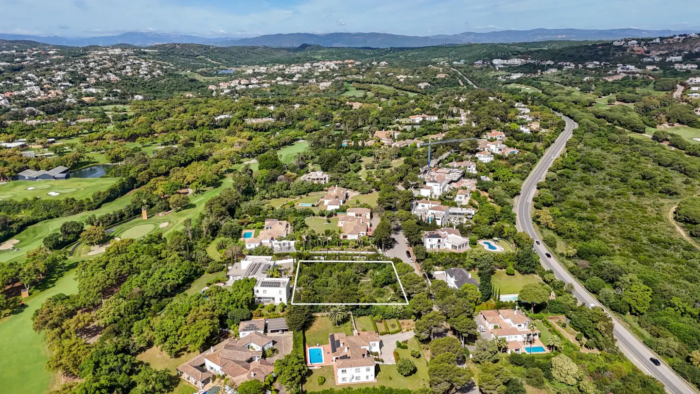 Aerial view of a property lot outlined in white, surrounded by lush greenery, houses, and a winding road. Mountains are visible in the distance.