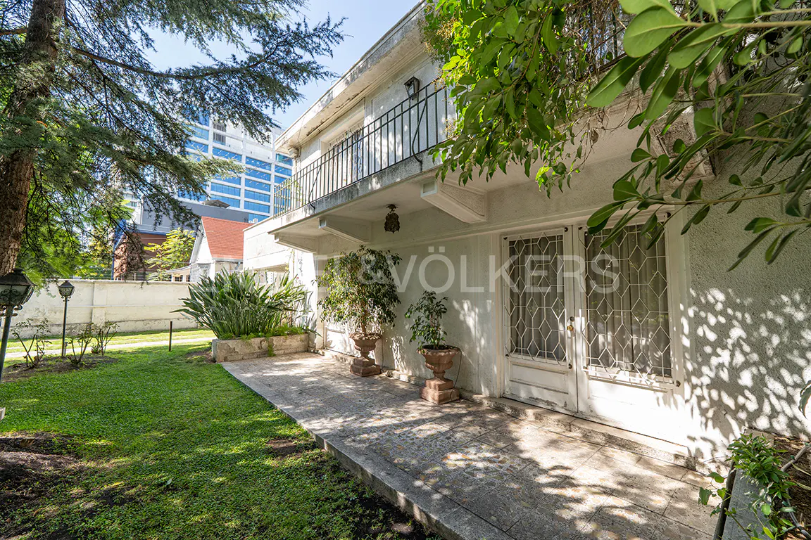 Exterior view of a two-story white house with a balcony, trees, and a green lawn. A modern building is visible in the background.