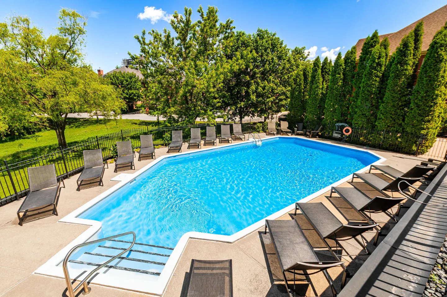 Outdoor swimming pool with bright blue water, surrounded by lounge chairs, lush green trees, and a black metal fence.