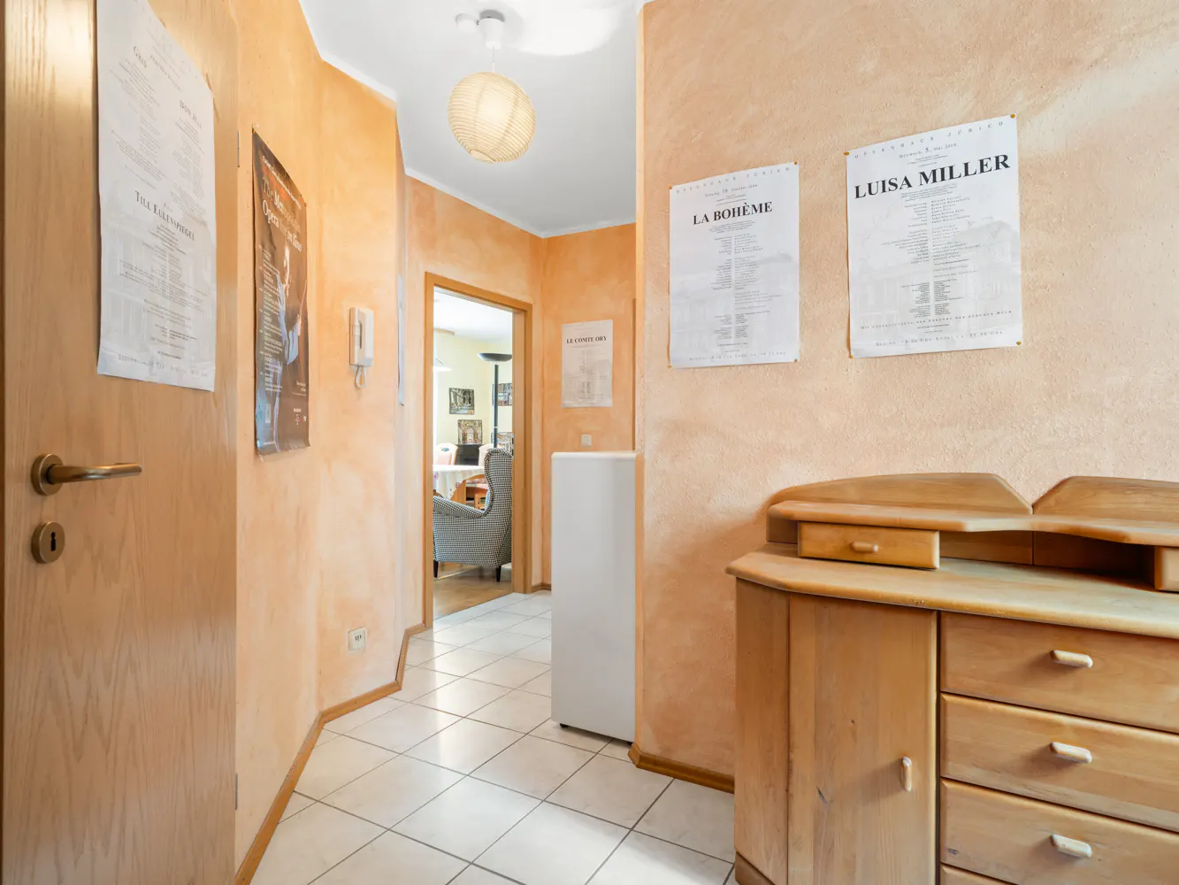Hallway with peach-colored walls, tiled floor, wooden dresser, and opera posters leading to a room with a grey armchair.