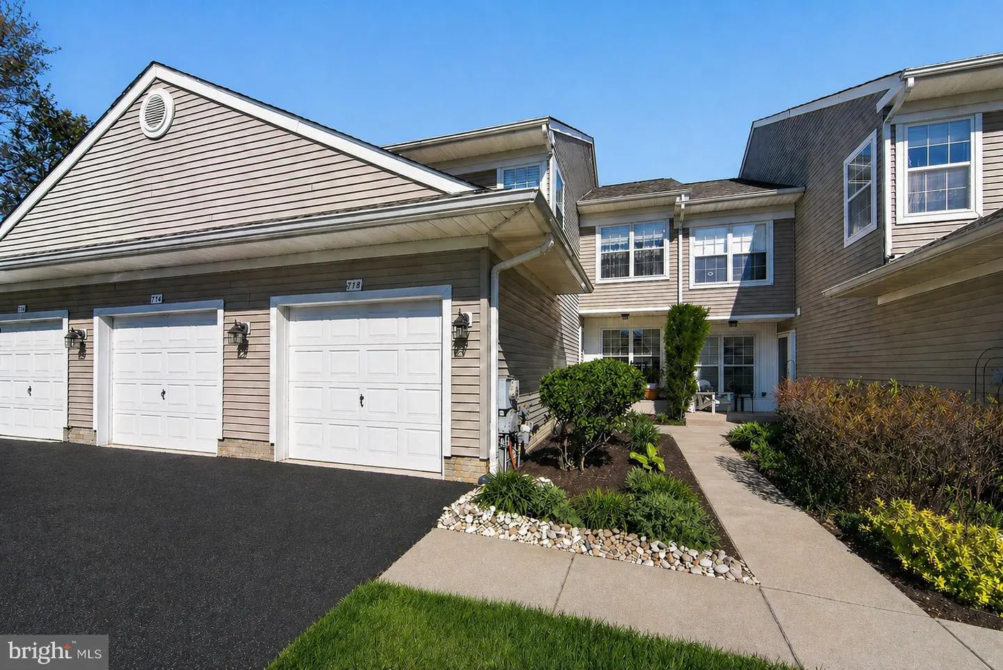 Tan two-story townhouses with white garage doors and a black asphalt driveway under a clear blue sky. A concrete walkway leads to the front doors.