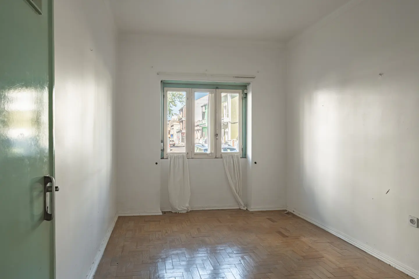 Empty white room with parquet floor, a window with white curtains, and a green door on the left.