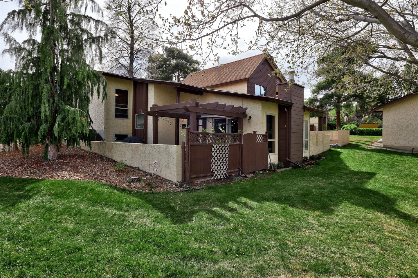 Backyard view of a beige townhouse with brown trim, a pergola, and a privacy fence. Green grass and trees surround the property.