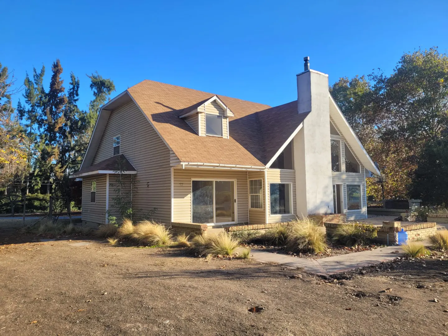 Tan two-story house with a brown roof, white chimney, and large windows under a clear blue sky.