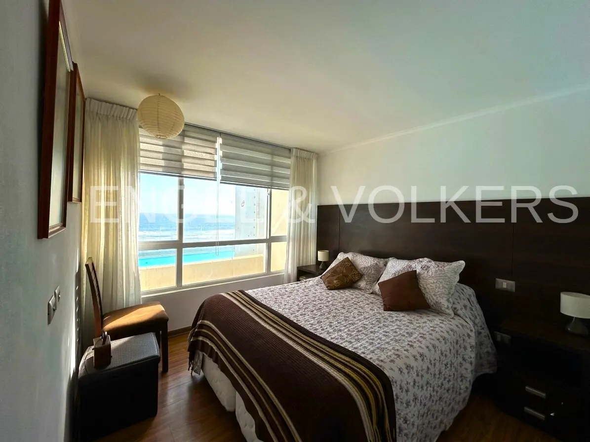 A bedroom with a large window overlooking the ocean. The bed has a brown and white patterned comforter. There are two nightstands and a dark wood headboard.