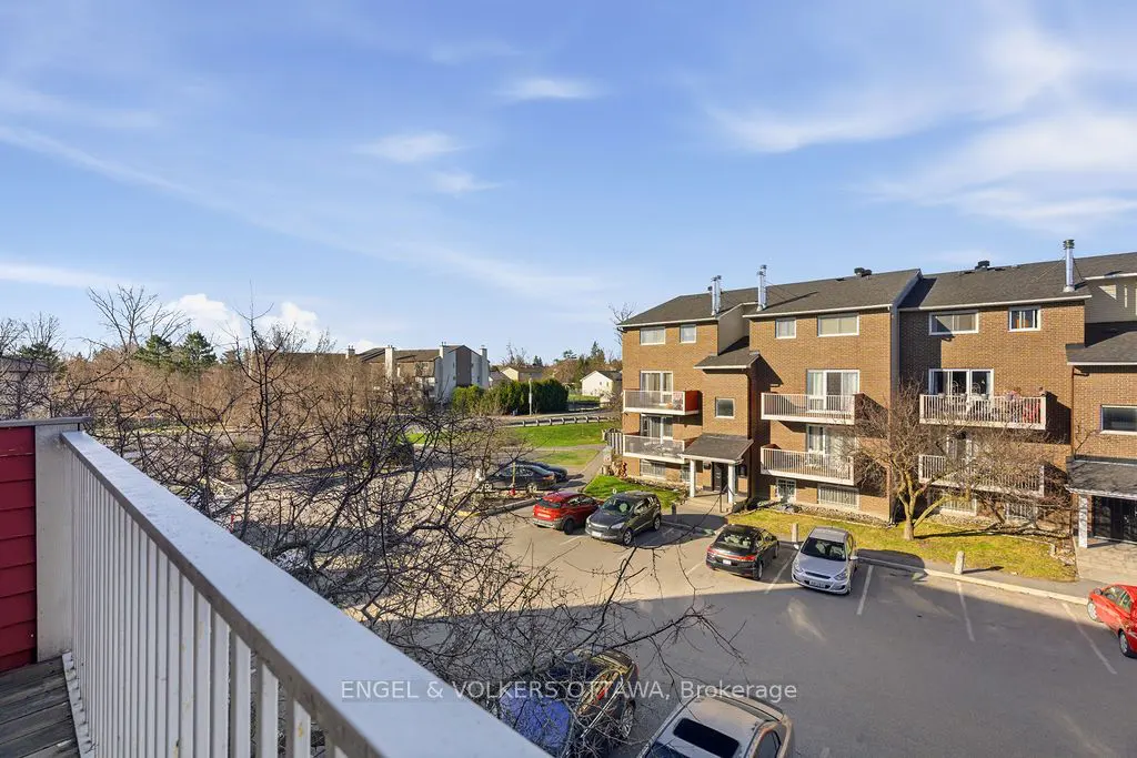 View from a balcony overlooking a parking lot with cars and a brick apartment building under a blue sky.