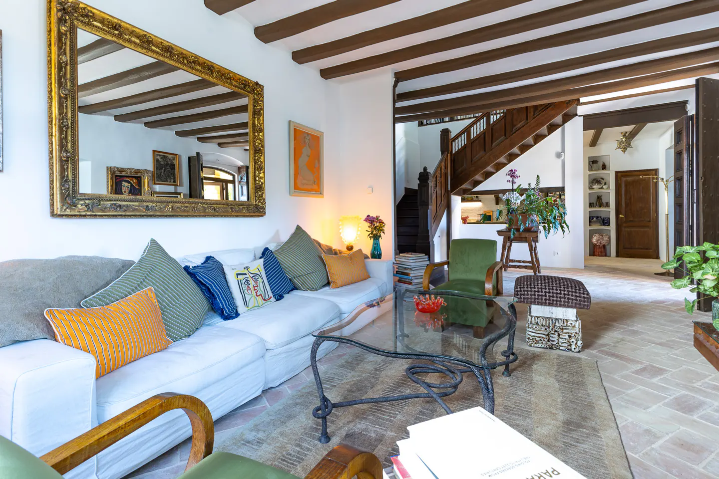 Living room with white sofa, colorful pillows, glass table, and wooden beams. A large gold-framed mirror hangs on the wall.