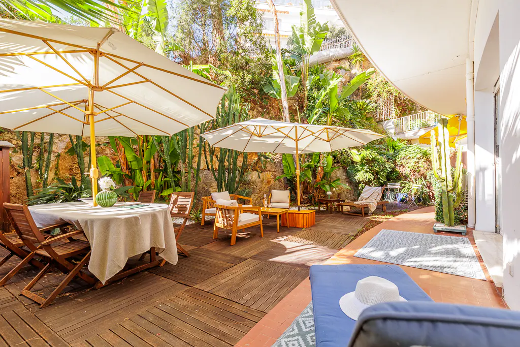 Outdoor patio with wooden deck, table, chairs, and lounge chairs. White umbrellas provide shade. Lush green plants and trees surround the area.